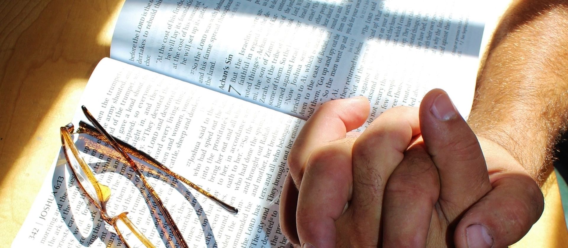 Hands clasped in prayer over an open Bible and reading glasses